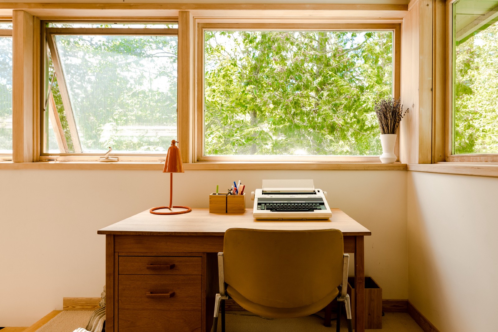a simple desk in a well lit room with a typewriter and big windows looking out on green trees