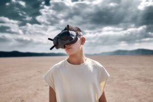 young man standing the desert in a white t shirt with a VR headset on, against a cloudy sky and distant desert mountains